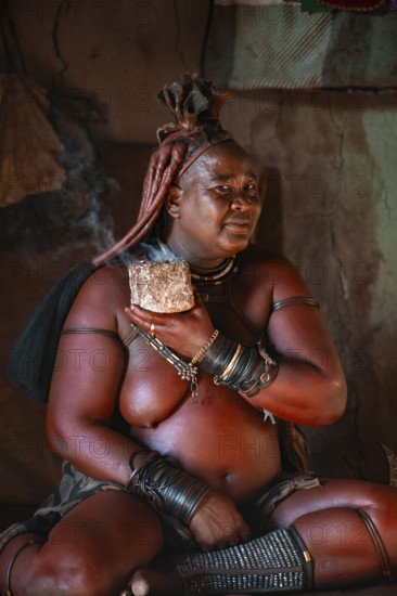 Natural perfume, elderly Himba woman smokes her hair for good smell, in a hut, traditional Himba