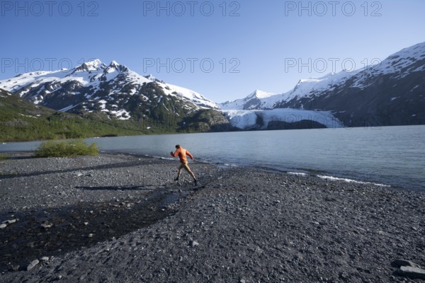 Young man jumping on a pebble beach on a glacial lake, Snowy Mountains and Portage Glacier on