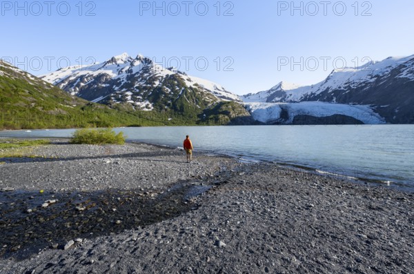 Young man on a pebble beach on a glacial lake, snowy mountains and Portage Glacier glacier on