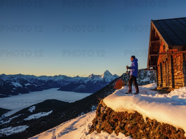 Mountaineers enjoy the sunrise in front of an alpine hut in winter, the Watzmann behind, fog in the