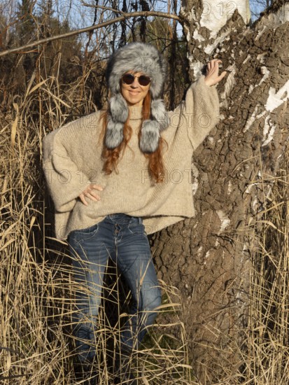 Slim woman with fur hat and sunglasses, red hair and warm winter colors between birch forest and
