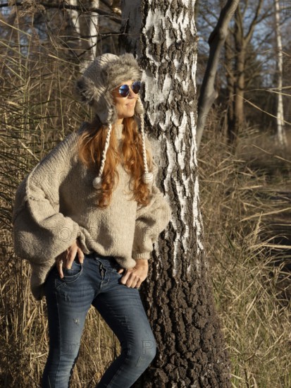 Slender pretty woman with fur hat and pompoms, red hair and warm winter colors between birch forest