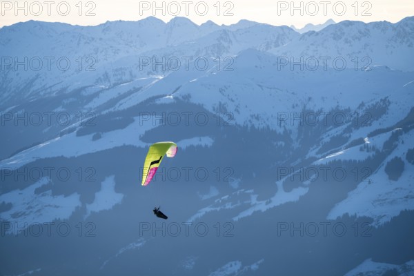 Paragliders flying over snowy mountain peaks in winter in evening light, Kitzbühel Alps, Tyrol,