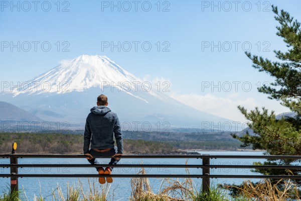 Young man sitting on railings next to a road and looking across the lake to Mt Fuji volcano, Motosu