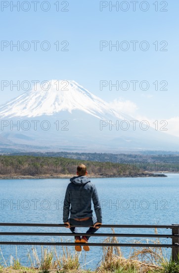 Young man sitting on railings next to a road and looking across the lake to Mt Fuji volcano, Motosu