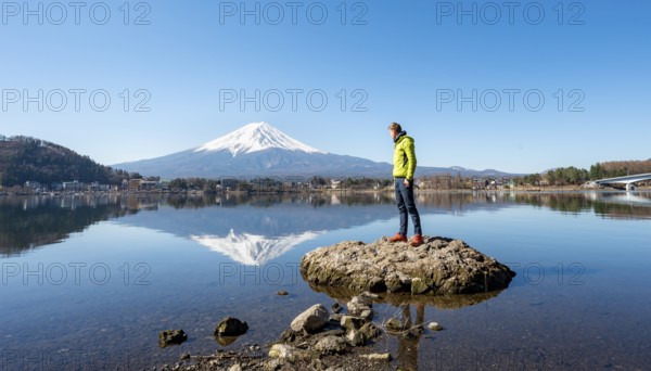 Young man standing on a rock in water, volcano Mt. Fuji is reflected in Lake Kawaguchi, Yamanashi