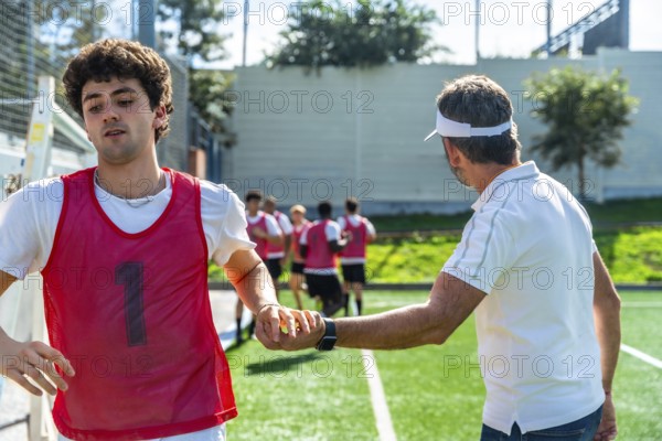 Coach instructing a soccer player on the field during training, teammates jogging in the background