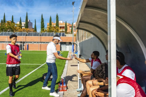 Soccer coach distributing water bottles to young male players sitting in the dugout during a