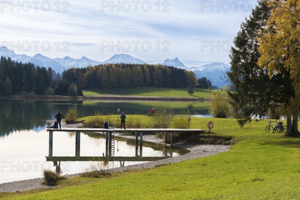 People walk along the jetty with views of Lake Forggensee and surrounding mountains, autumn trees