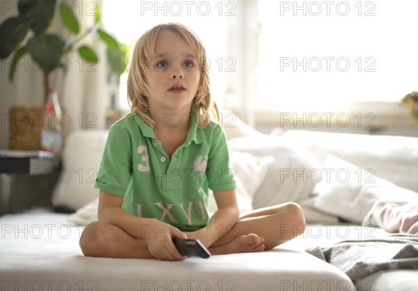 Child, boy, 7 years old, blonde, sitting on sofa, with remote control in hand, watching TV,