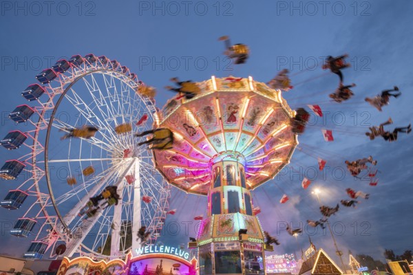 An illuminated chain carousel and Ferris wheel at night at a funfair, people swinging on chains,