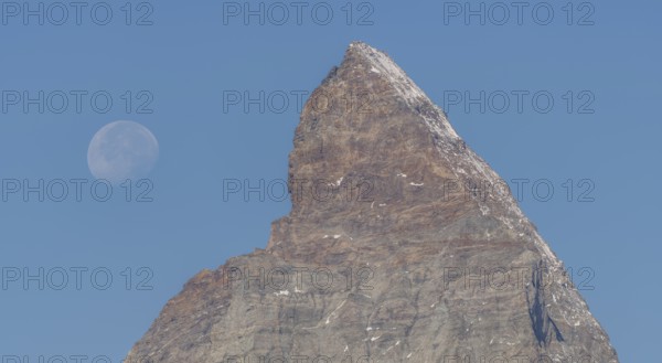 The Matterhorn mountain peak rises steeply against a bright blue sky. The full moon glistens gently