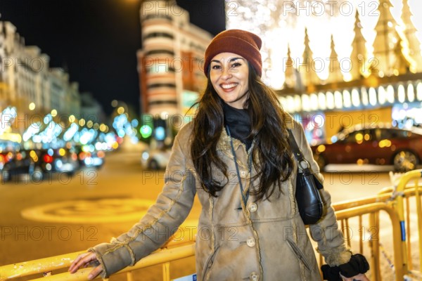 Young woman wearing winter clothes smiling in a city street decorated with christmas lights at