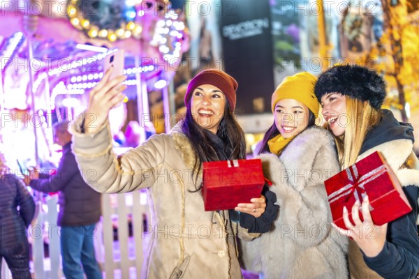 Three happy women holding christmas gifts are taking a selfie in front of a carousel in a christmas
