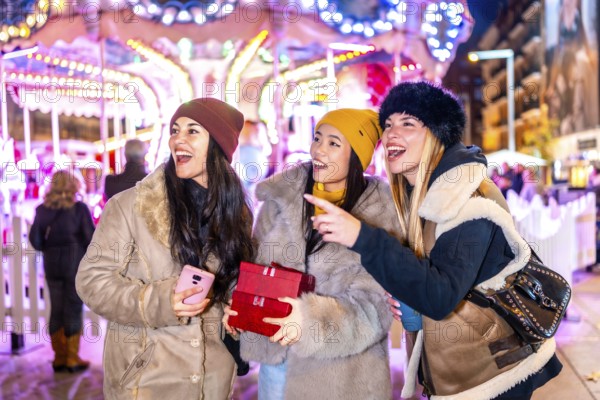 Three happy female tourists holding christmas gifts pointing at something at the christmas market