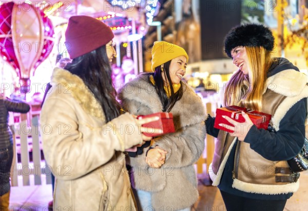 Three cheerful female friends exchanging christmas gifts at a night market with illuminated