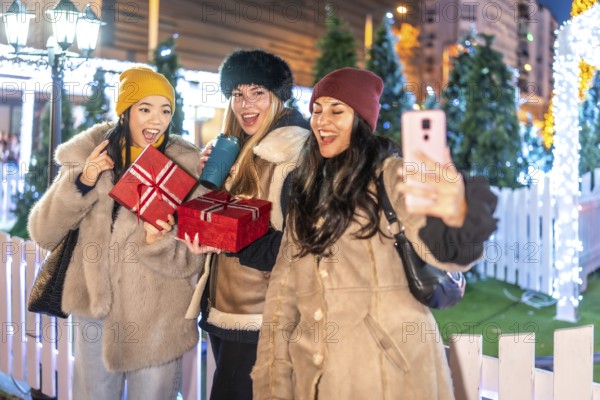 Three cheerful female friends holding christmas gifts and taking a selfie at a decorated outdoor