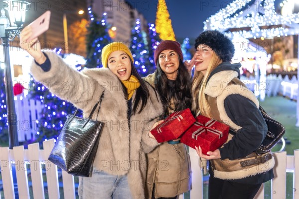 Three cheerful women taking selfie with christmas gifts and sticking out tongue in christmas market
