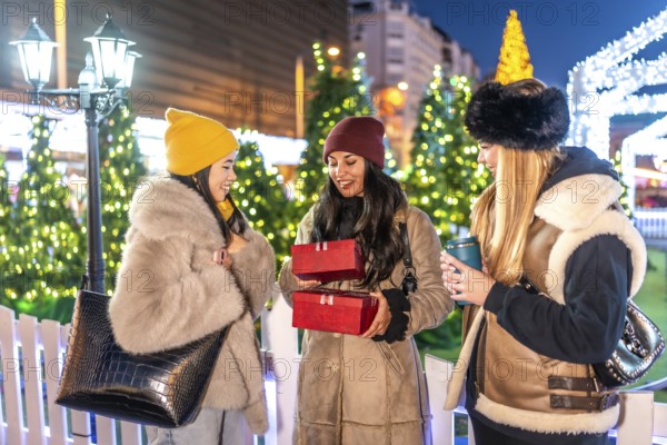 Three friends exchanging christmas presents and enjoying hot drinks at a decorated outdoor