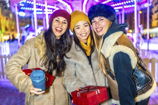 Three happy female friends enjoying christmas market at night, holding gifts and hot beverages,