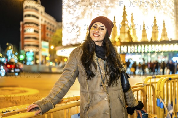 Smiling young woman leaning on a fence in a city street enjoying christmas lights and decorations