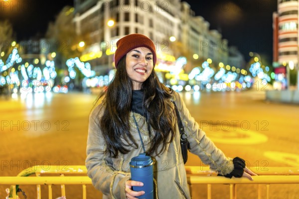 Young woman wearing winter clothes, holding a thermal mug and smiling near a street fence with