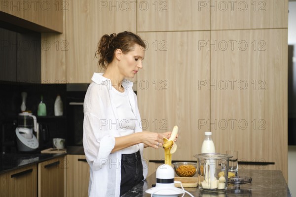 In a stylish kitchen, a woman is focused on preparing a smoothie by peeling a banana. Nearby, a