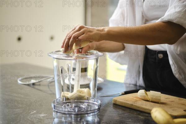 Sliced bananas are placed in a blender while a sunny kitchen provides a bright backdrop. Fresh