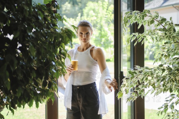A young woman stands at the entrance of a bright garden space, holding a glass of iced beverage.