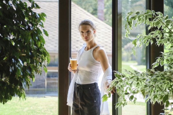 A woman stands by a large window, sipping a cool beverage while surrounded by green plants and