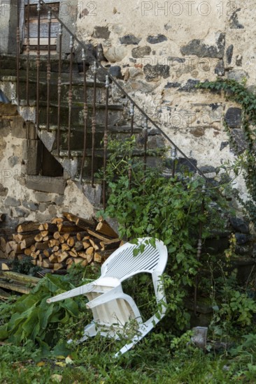 An unkempt outdoor area features a white chair leaning against a weathered stone wall, surrounded