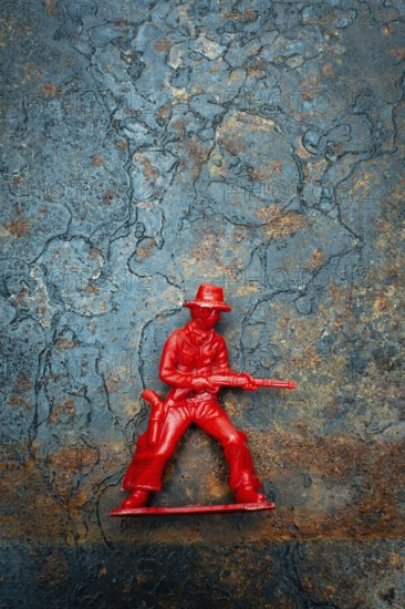A small red plastic toy cowboy stands firmly on a rustic blue and brown textured background. The