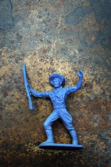A small blue plastic toy cowboy stands firmly on a rustic blue and brown textured background. The