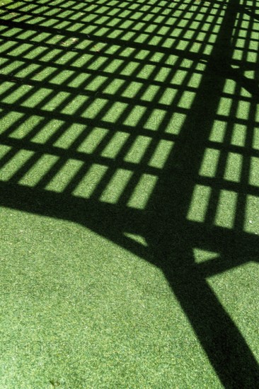 A grid of shadows falls across vibrant green turf, formed by a lattice overhead. The sunlight