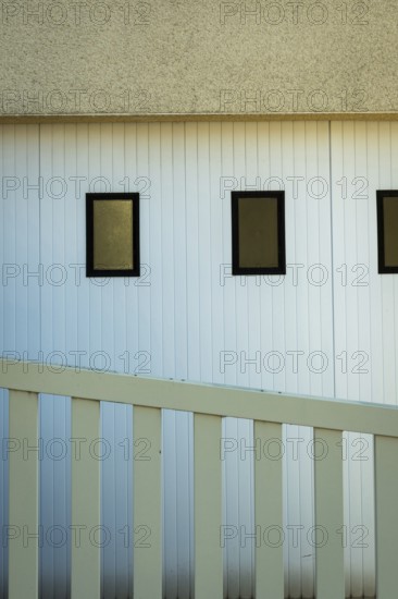 A building featuring three rectangular windows on a white vertical paneled wall. A white railing