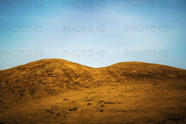 Two gentle hills rise from a golden landscape under a bright blue sky. Wispy clouds streak across