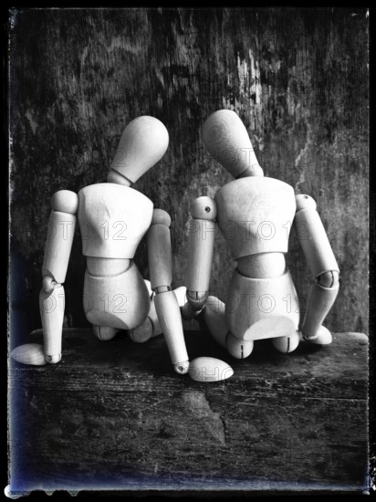 Two wooden figures are positioned closely together, facing away from the viewer. They sit on a