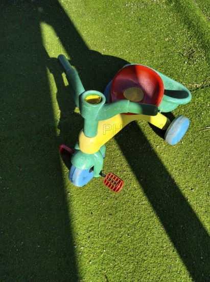 A vibrant, multi-colored tricycle sits on lush, green artificial grass, casting a shadow in bright