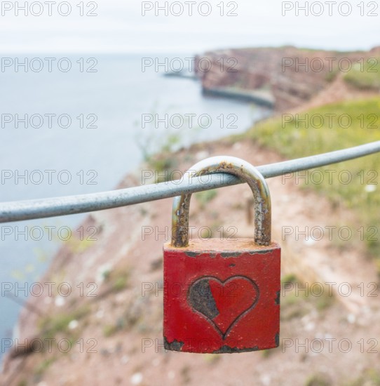Red padlock with engraved heart symbol on a wire rope in front of the steep coast, red cliff of