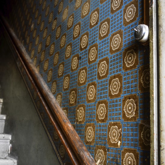 A close-up view of old wallpaper lining the walls of a staircase corridor, featuring a geometric