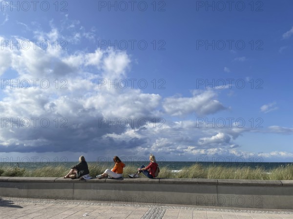 Three holidaymakers enjoying the sun, sitting on a little wall on the promenade, blue sky, Baltic
