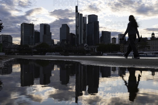 A woman walks along the banks of the Main in the rain in front of the Frankfurt banking skyline,