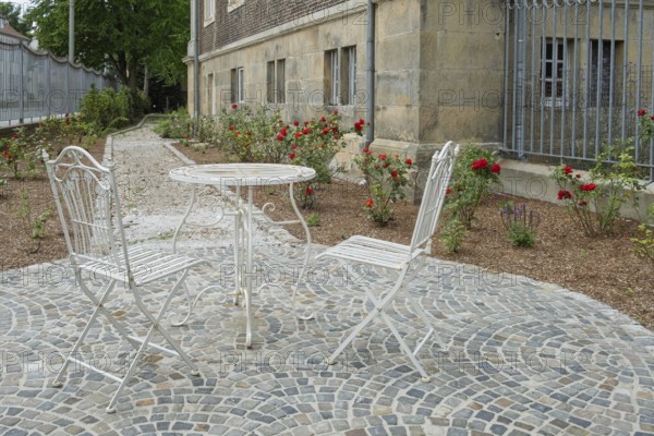Two white metal chairs and a table on a paved square in front of a historic building, surrounded by