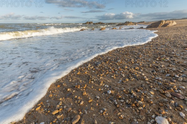 A wide sandy beach stretches out with scattered pebbles and beach shells. Gentle waves crash