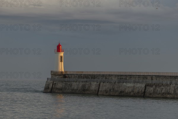 A lighthouse stands on a pier and casts its light over calm waters at dusk. The sky is filled with
