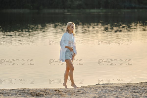 A woman strolls barefoot on a sandy beach by the lake, enjoying the serene atmosphere as the golden