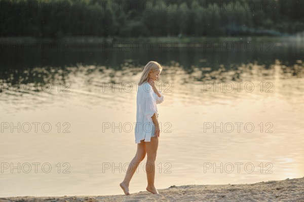 Pensive woman strolls barefoot on the sandy beach beside a serene lake. Golden sunlight reflects