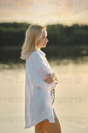 A young woman stands pensively by a calm lake at sunset, dressed in a light shirt and shorts. The