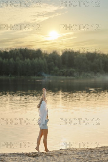 A woman stands on the sandy shore of a tranquil lake, raising her hand joyfully towards the setting