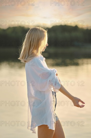 A young woman stands by the tranquil lake at sunset, wearing a white shirt and denim shorts. The